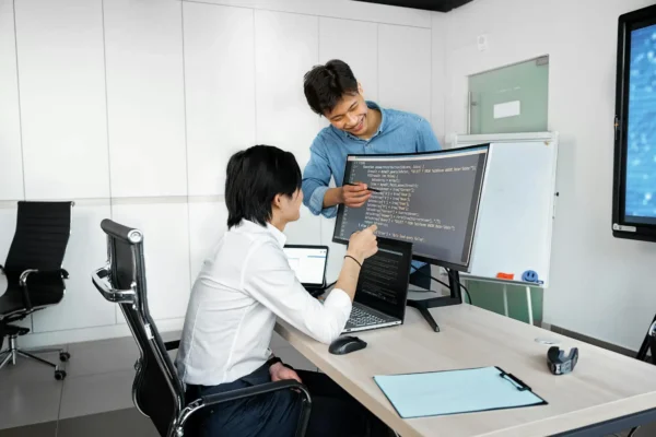 Two IT interns collaborate in an office, discussing code displayed on a large monitor, while one points to the screen and the other engages from a seated position.