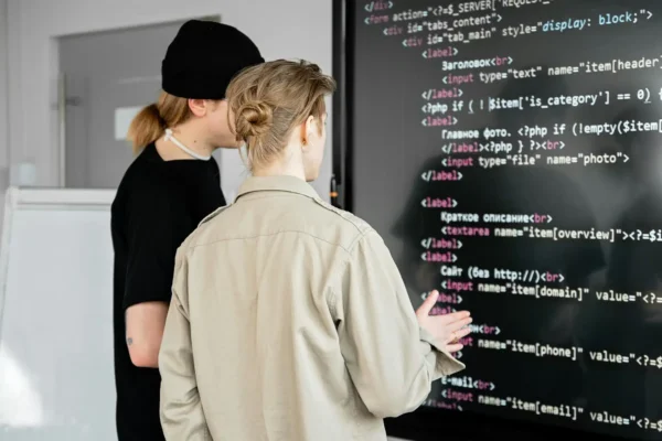 Two IT interns stand closely in front of a large screen filled with code, reviewing and discussing the programming details displayed.