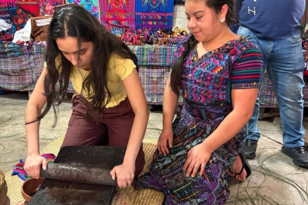 An intern grinds cacao on a traditional metate, guided by an indigenous woman in a vibrant cultural market.