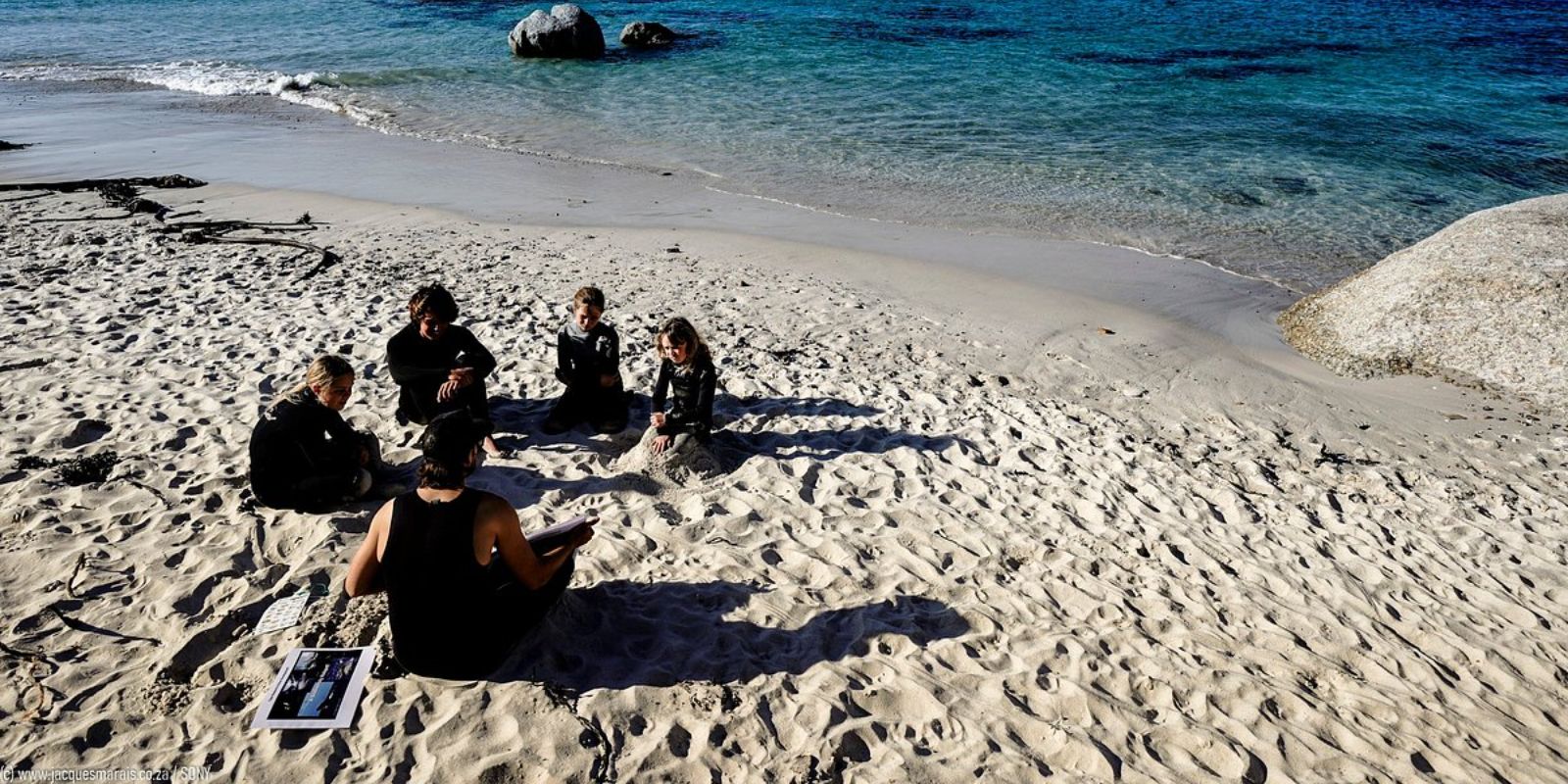 A small group sits on sand near the shoreline, having a lesson by the sea