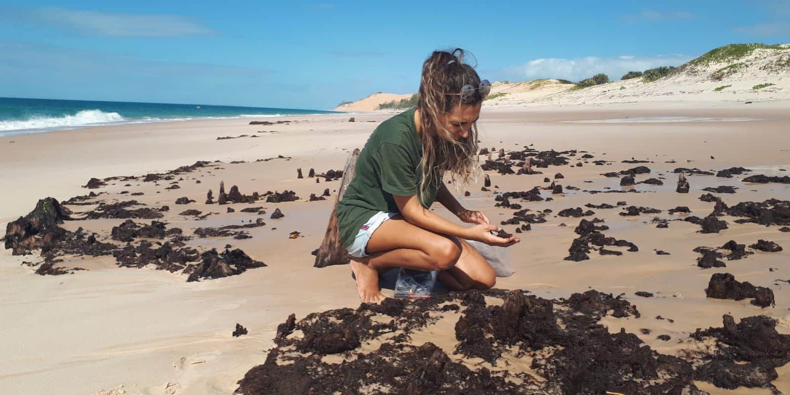 A woman kneels on sandy shore examining seaweed washed up on the beach