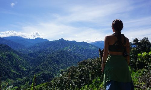 Person standing on a hilltop, looking out over green mountains under a bright blue sky
