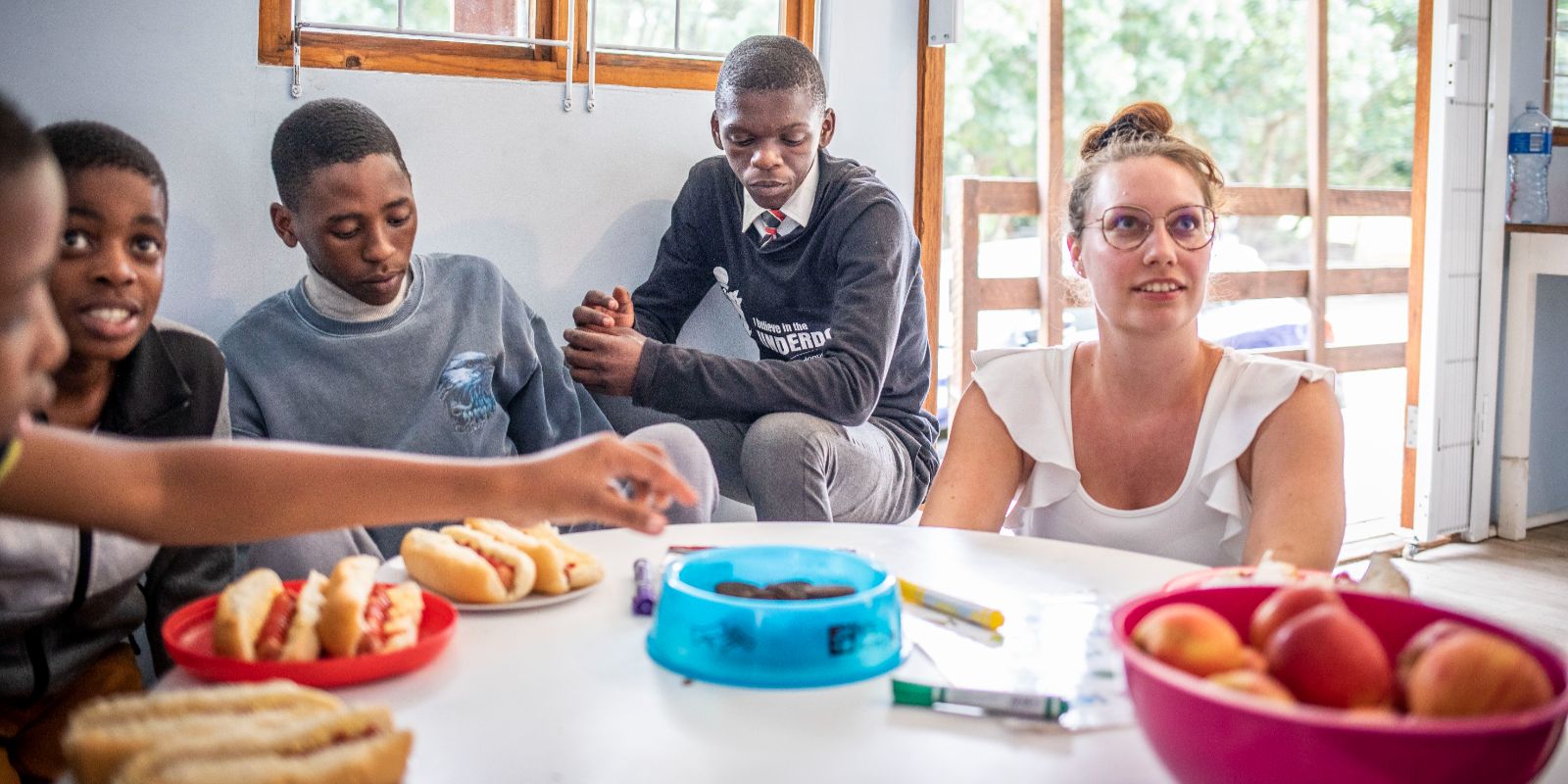Group of young people and a woman sitting at a table with hot dogs and fruit