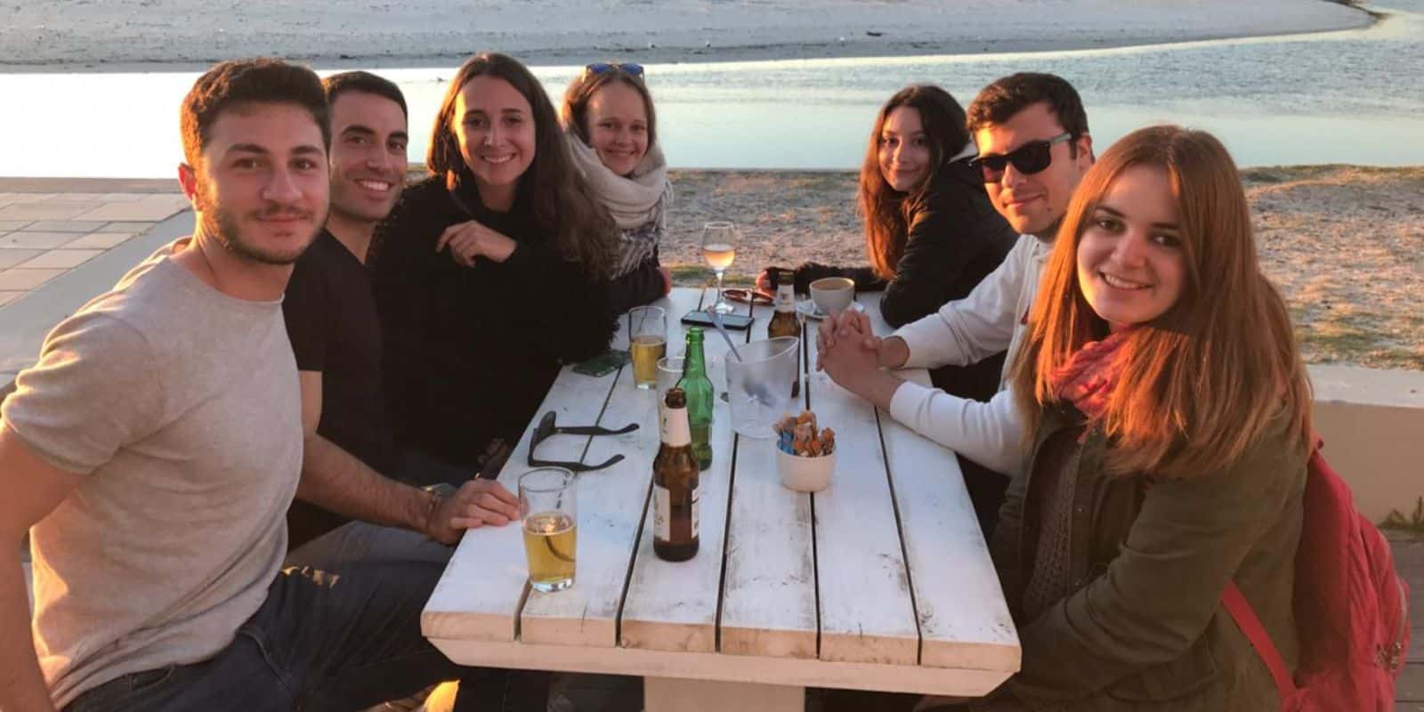 Friends sitting at a picnic table by the beach at sunset, smiling at the camera