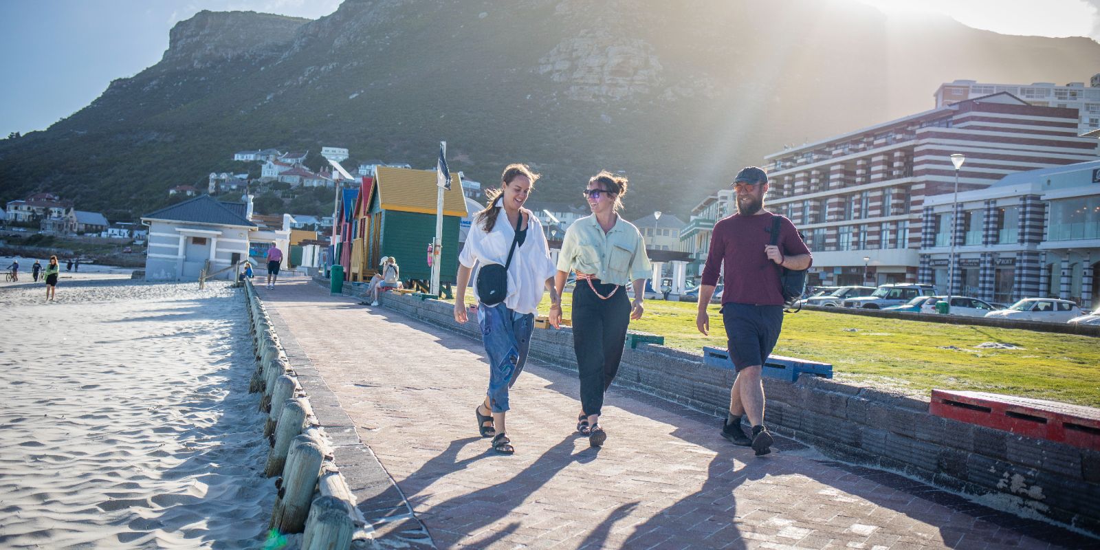 Three people walking along a beachside path with colorful huts and mountains in the background