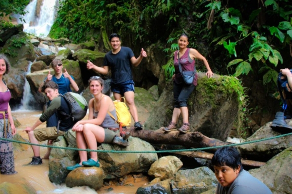 Group of people standing and sitting on rocks near a small waterfall in a forested area