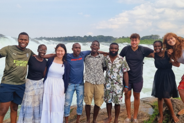 A group of nine people posing in front of a body of water. Their arms are around one another.