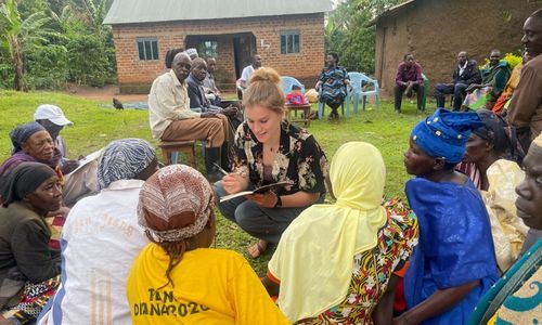 An intern crouching and speaking to people sitting on the ground while writing in a notebook.