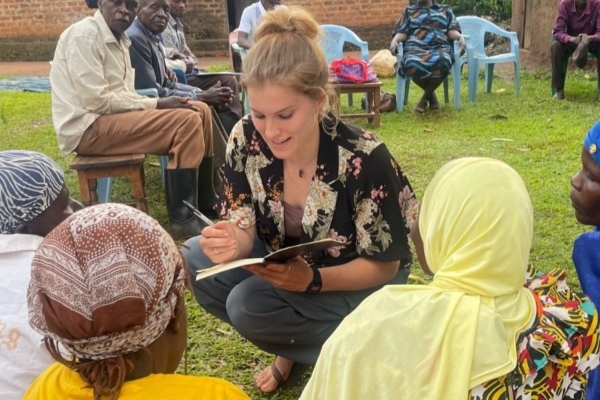 An intern crouching and talking to people sitting on the ground while taking notes in a notebook.